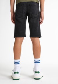 Bermudas de mezclilla negras con corte recto, con puños enrollados y bolsillos traseros, combinadas con calcetas blancas con rayas azules y zapatillas con acentos verdes.