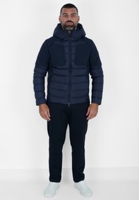 Giacca puffer blu navy con zip frontale, cappuccio e due tasche sul petto; presenta un design a righe orizzontali; abbinata a pantaloni scuri e sneakers bianche.