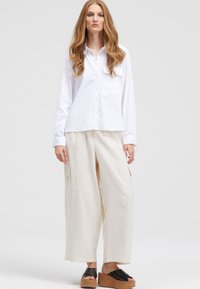 White long-sleeve button-up shirt with a single chest pocket, paired with loose-fitting, cream-colored trousers. Model wears black platform sandals.