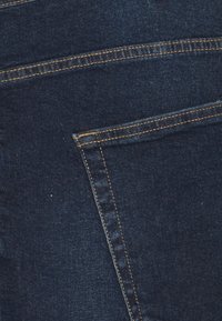 Tela de mezclilla azul oscuro con una textura suave. Presenta costuras visibles en un color dorado contrastante y un detalle simple de bolsillo trasero.