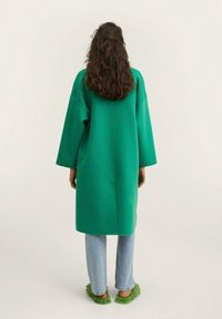 Grønt oversized frakke med sænkede skuldre. Blød tekstur, lige snit. Sammen med lyseblå jeans og fluffy grønne slippers.