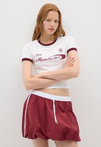 Camiseta blanca corta con acentos en color burdeos y texto "Manchester", combinada con pantalones cortos sueltos en color burdeos que cuentan con un cordón blanco y rayas laterales.