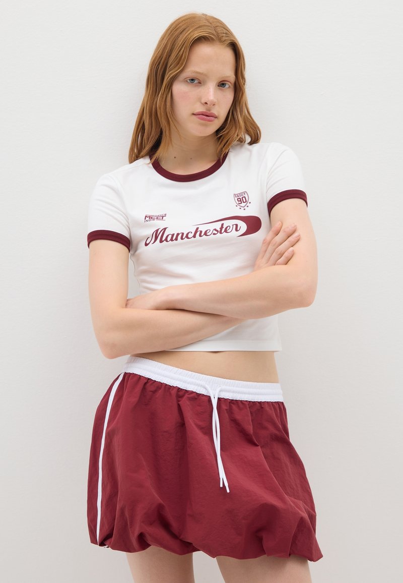 Camiseta blanca corta con acentos en color burdeos y texto "Manchester", combinada con pantalones cortos sueltos en color burdeos que cuentan con un cordón blanco y rayas laterales.