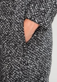 Tekstureret sort og hvid ulden frakke med en lomme i siden, der viser et vævet mønster med en let chunky finish.