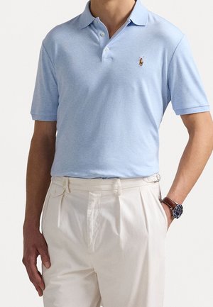 Mann trägt ein hellblaues Poloshirt mit kleinem Logo, cremefarbene plissierte Hose und eine braune Armbanduhr, Hand in der Tasche.