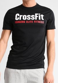 Černé tričko s krátkým rukávem s velkým bílým nápisem "CrossFit" a červeným nápisem "FORGING ELITE FITNESS" na přední straně. Měkká tkanina, přiléhavý střih.