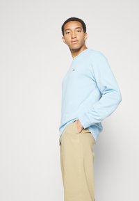 Sweatshirt bleu clair à manches longues, avec un col rond simple et un petit logo sur la poitrine, assorti à un pantalon beige.