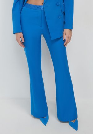 Blue flared trousers made of smooth fabric, featuring a fitted waist and slanted pockets, paired with pointed blue heels.