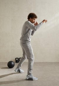 Szary sportowy dres z prążkowanymi detalami i paskowymi akcentami; o luźnym kroju, zestawiony z szarymi sneakersami. W tle sprzęt do fitnessu.