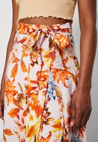 Žena v béžovém zkráceném topu a sukni s vysokým pasem, na které je oranžový, hnědý, žlutý a modrý květinový vzor uvázaný na mašličku v pase.