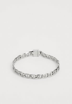 Brățară din metal de culoare argintie, cu închidere rectangulară gravată cu numele mărcii, așezată plat pe un fundal alb simplu.