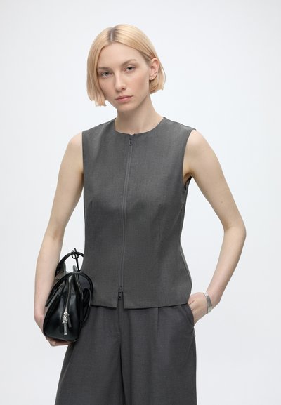 Femme aux cheveux blonds courts portant un haut gris sans manches et un pantalon assorti, tenant un sac à main noir dans une main.