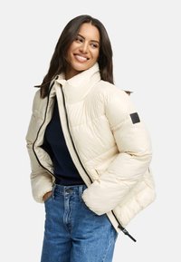 Polstret cremet pufferjakke med høj krave, sort lynlås og logopatch. Bæret åben, parret med en marineblå top og blå jeans.