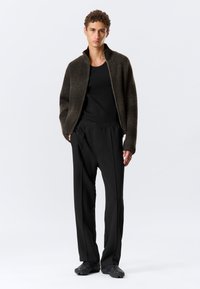 Rjava rebrasta jakna na zadrgo, črna majica brez rokavov, črne ohlapne hlače in črne teksturirane čevlje z edinstvenimi detajli.