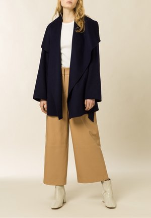 Personne portant un manteau oversize bleu marine, une chemise blanche, un pantalon beige large et des bottines blanches, debout devant un fond clair uni.