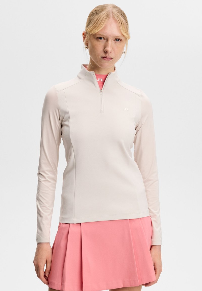 Blonde vrouw die een beige top met lange mouwen en een kwart-ritssluiting draagt, en een roze geplooide rok, staand tegen een effen lichte achtergrond.