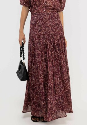 Mujer con una falda larga floral color granate y una blusa con mangas abullonadas, sosteniendo un bolso negro con asa de cuentas, posando frente a un fondo liso.