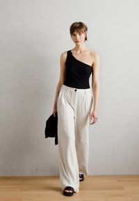 Schwarzer einseitiger Bodysuit, weiß gestreifte weite Hose, schwarze Sandalen. Model hält eine schwarze Jacke in einem minimalistischen Interieur.