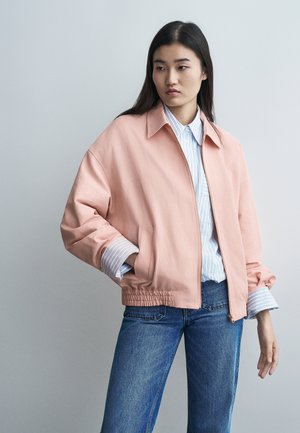 Fiatal nő világos rózsaszín dzsekit visel, kék-fehér csíkos inggel és kék farmerrel, egyszerű világosszürke háttér előtt áll.