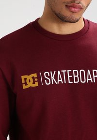 Maroon sweatshirt i bomull med en gul DC-logotyp och vit "SKATEBOARD" text framtill. Ribbad halsringning, slät struktur.
