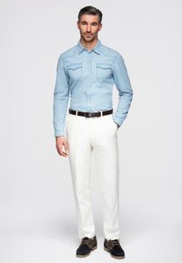 Chemise en denim bleu clair avec deux poches poitrine, pantalons blancs et chaussures foncées avec des accents marron. Le modèle se tient sur un fond neutre.