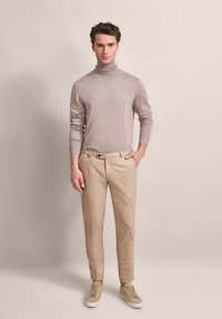 Helles grauer Rollkragenpullover, beige maßgeschneiderte Hose und cremefarbene Sneaker. Glatte Textur, figurbetontes Design und dezente Rippung am Pullover.