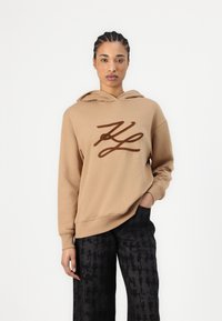 Hanorac cu glugă maro, având un logo mare, de culoare maro închis. Textură moale, croială lejeră. Asortat cu pantaloni negri cu un model, având un design lejer.