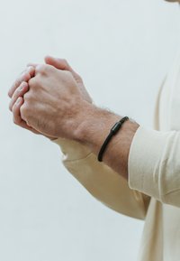 Pulsera de cuero trenzado negro con un cierre metálico cilíndrico, llevada en una muñeca, manos juntas, sobre un fondo neutro.