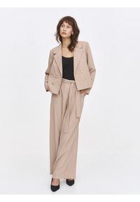 Beige double-breasted blazer with gold buttons, paired with a black top and matching beige trousers featuring a tie belt and wide legs. Black heels.
