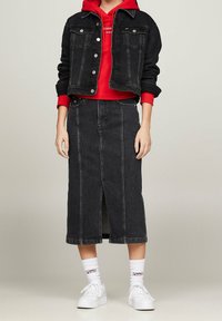 Personne portant un sweat à capuche rouge sous une veste en denim noir raccourcie, une jupe en denim noire midi avec une fente à l'avant, des chaussettes blanches et des baskets blanches.