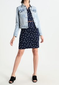 Hellblaue Jeansjacke über einem marineblauen Kleid mit Polka-Dots und taillierter Silhouette. Schwarze, offene Mules mit lässigem Design und strukturierter Oberfläche.