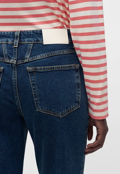 Personne portant un jean bleu avec des poches arrière et un shirt à manches longues rayé rose et blanc, main détendue le long du corps.