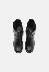 Une paire de bottines noires en cuir avec des bouts carrés arrondis et des sangles à boucle sur le côté, vue de dessus sur un fond blanc.