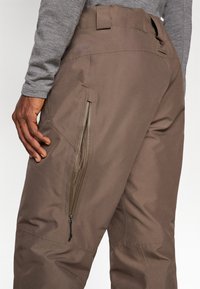 Brown waterproof pants with a smooth texture, featuring side zip pockets and a fitted waist. Reinforced stitching showcases durability.