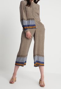 Brun mönstrad jumpsuit med långa ärmar, blå och orange detaljer vid muddar och fåll, samt kort vid benet design. Buren med beige klackskor med blockklack.