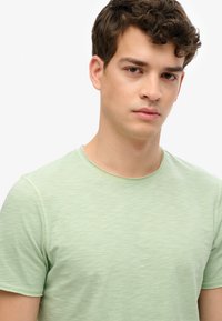 Camiseta de manga corta verde claro con escote redondo, confeccionada en una suave tela texturizada, que presenta un sutil patrón desgastado a lo largo de toda la prenda.