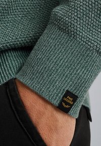 Manche de pull en tricot vert foncé avec un motif texturé ; présente une étiquette noire avec "PME LEGEND" en doré ; détail de poignet côtelé.