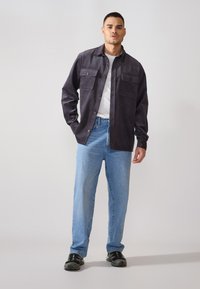 Chemise-veston en velours côtelé gris foncé avec deux poches poitrine, portée sur un t-shirt blanc et assortie à un jean bleu clair et des baskets noires.