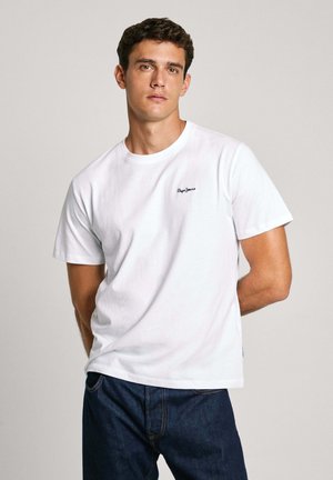Camiseta de algodón blanca con cuello redondo, mangas cortas y un pequeño logo negro en el pecho. Llevada con jeans de mezclilla azul oscuro.