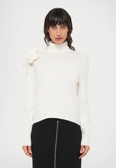 White turtleneck sweater with a side flower accent, made of a smooth fabric. The fit is relaxed with a ribbed hem. Paired with a black skirt featuring a gold zipper.