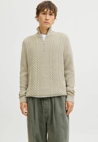Pull en maille zippé beige clair avec un motif textur�é, doté de poignets et d'un ourlet côtelés. Porté par-dessus une chemise blanche à col rond et associé à un pantalon olive.