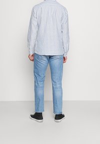 Homme portant une chemise à manches longues rayée bleu clair, un jean bleu clair et des baskets Converse noires, debout face à un mur blanc uni.