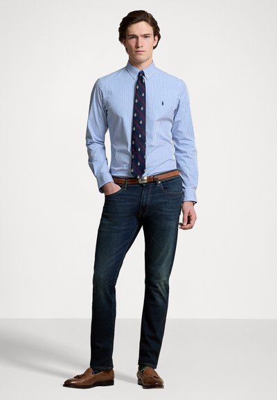 Camicia a righe blu e bianche con bottoni, cravatta blu scuro con accenti a motivo, cintura in pelle marrone, jeans di denim scuri, scarpe classiche marroni.