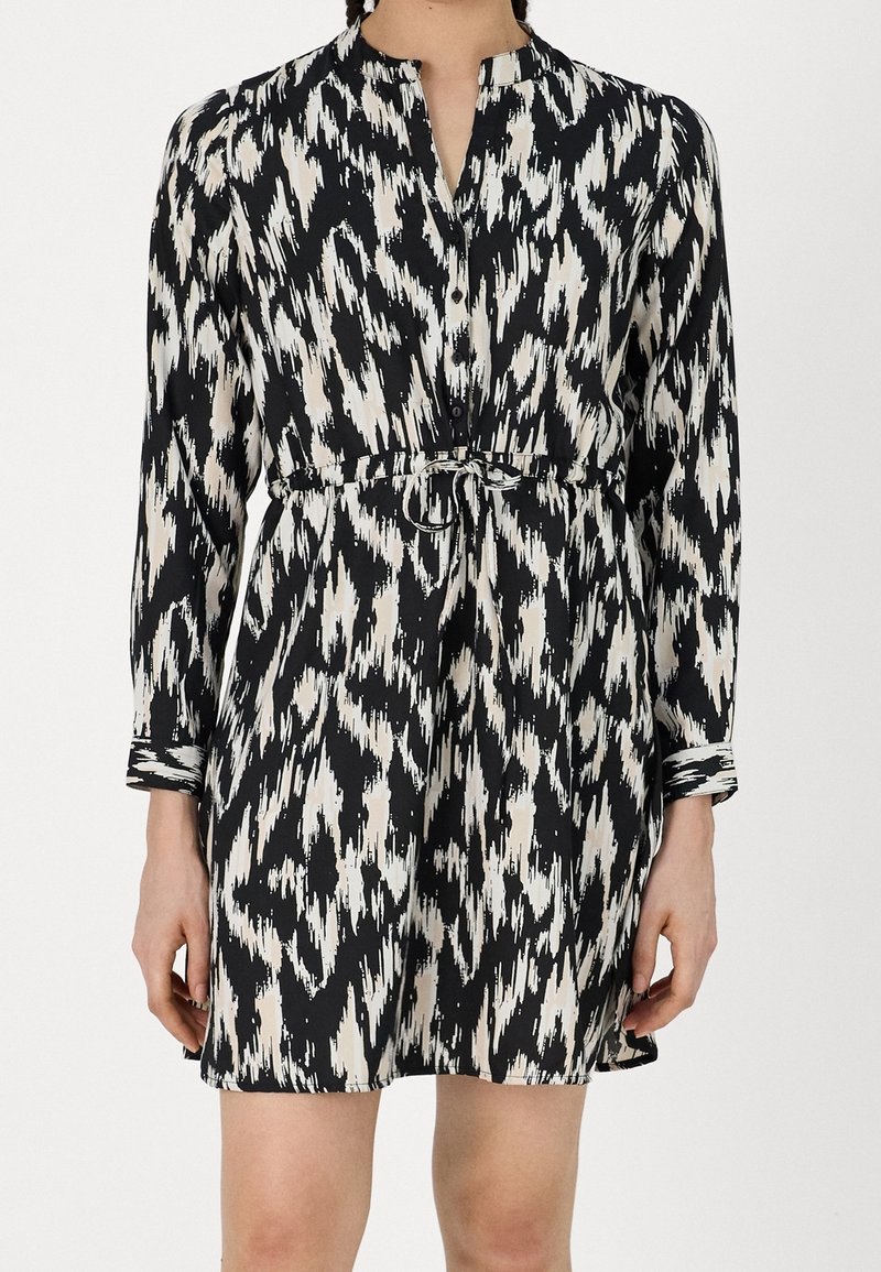 Robe à manches longues à motif abstrait en noir et blanc, avec une fermeture boutonnée sur le devant et une ceinture à la taille, portée par une personne avec les bras détendus le long du corps.