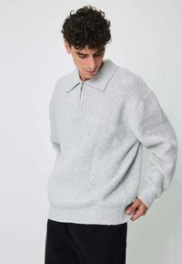 Pull-over gris clair texturé avec un col zippé, manches longues et coupe ample. Design simple et minimaliste avec des poignets côtelés.
