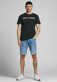 Schwarzes Baumwoll-T-Shirt mit weißem Logo, hellblaue Denim-Shorts mit umgeschlagenem Saum, kombiniert mit weißen Sneakern und sichtbaren Bein-Tattoos.