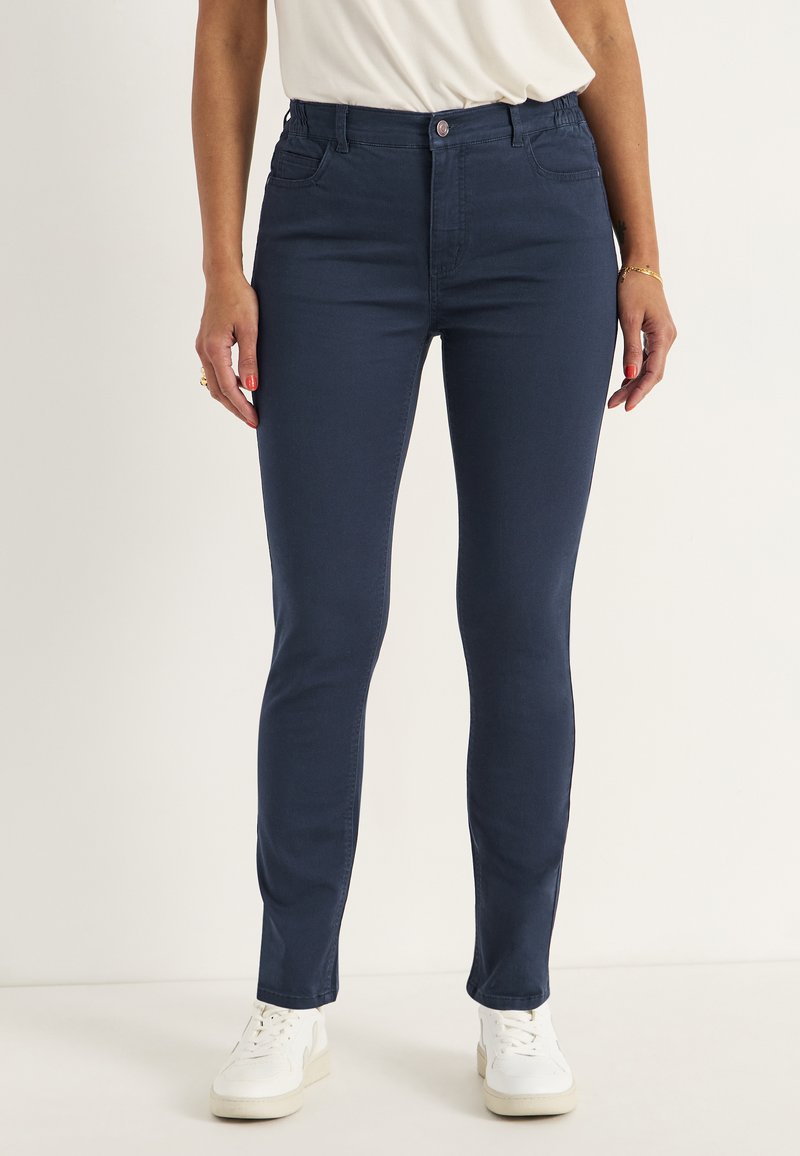 Marinblå stretchjeans med slim fit, medelhög midja, fem fickor och knappstängning, kombinerade med vita sneakers.