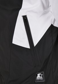 Cazadora negra y blanca con bolsillo con cremallera, diseño de bloques de color angular y etiqueta de marca cosida en la parte inferior derecha.