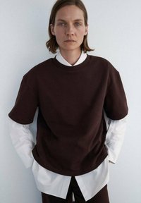 Brun oversized sweatshirt med korte ærmer, lagd over en skarp hvid skjorte, mod en ensfarvet baggrund, der viser et moderne, minimalistisk design.
