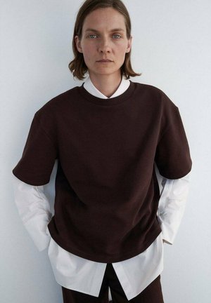 Brun oversized sweatshirt med korte ærmer, lagd over en skarp hvid skjorte, mod en ensfarvet baggrund, der viser et moderne, minimalistisk design.
